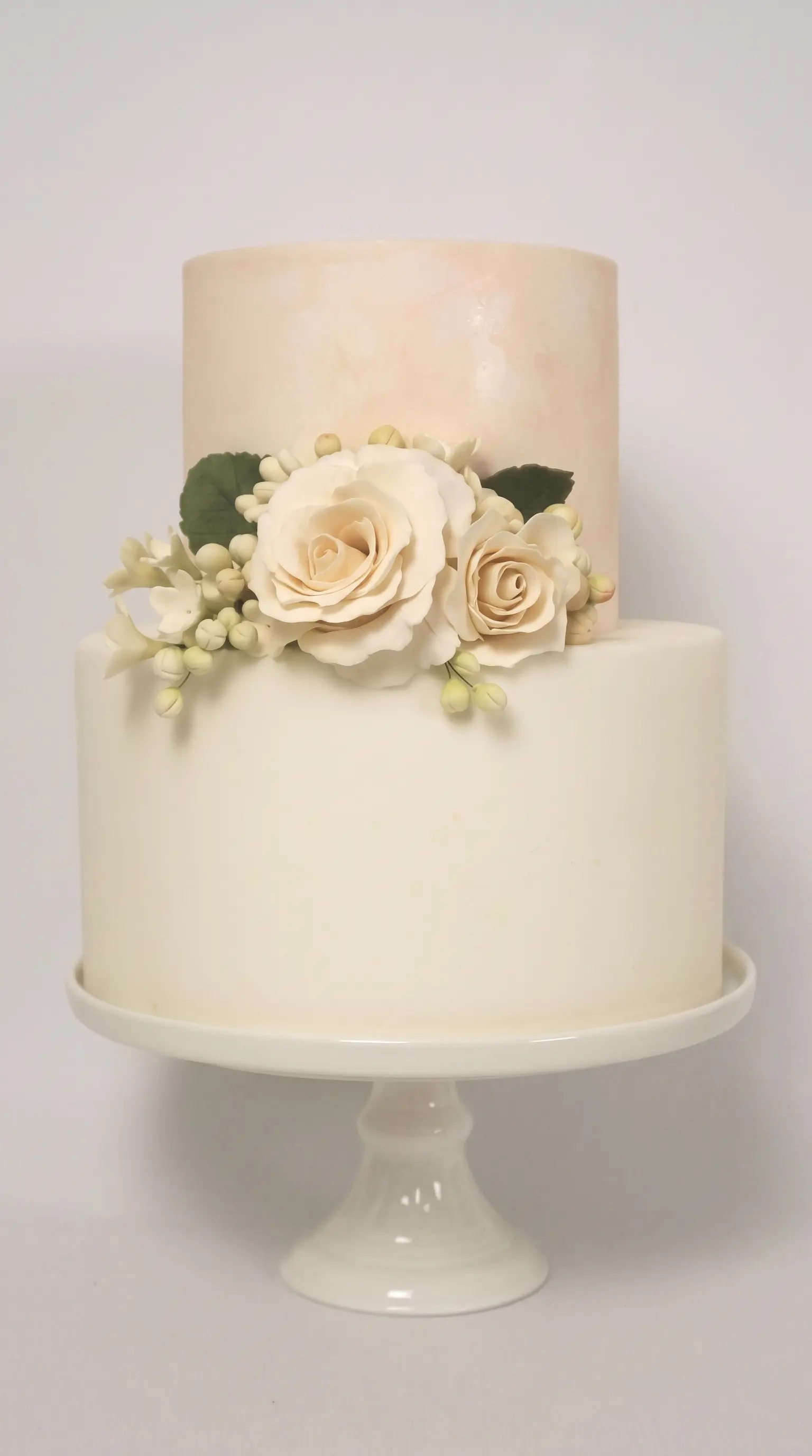 Ivory two-tier wedding cake decorated with delicate sugar flowers on a white cake stand